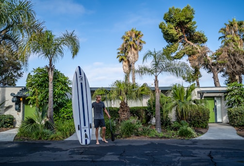 A person stands barefoot on a black paved surface holding a paddle next to a large white surfboard with blue stripes. The background features a single-story building with green doors, surrounded by lush green palm trees and various foliage. The sky is clear with some clouds, suggesting a sunny day.