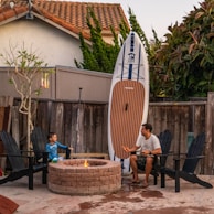 A man and a child are seated on black patio chairs around a small round fire pit in an outdoor area. Behind them is a wooden fence, and a large paddleboard is propped up against it. Nearby, there is a leafless tree with hanging flowers and a bush with reddish leaves. The scene is set in a suburban backyard with a tiled roof house in the background.