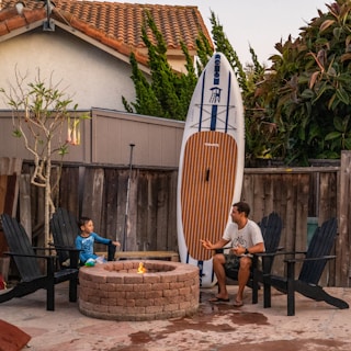 A man and a child are seated on black patio chairs around a small round fire pit in an outdoor area. Behind them is a wooden fence, and a large paddleboard is propped up against it. Nearby, there is a leafless tree with hanging flowers and a bush with reddish leaves. The scene is set in a suburban backyard with a tiled roof house in the background.