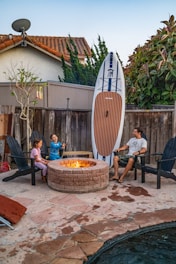 A family scene in a backyard features a man and two children seated around a stone fire pit. The children are sitting on Adirondack chairs, with one holding a roasting stick. Leaned against the wooden fence is a large paddleboard. The setting includes a small tree, a satellite dish on the rooftop of a nearby house, and lush greenery surrounding the area.