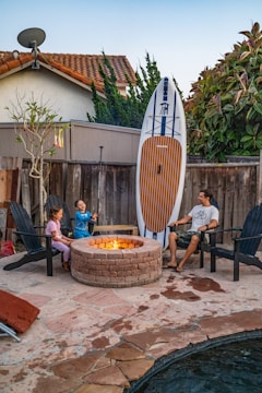 A family scene in a backyard features a man and two children seated around a stone fire pit. The children are sitting on Adirondack chairs, with one holding a roasting stick. Leaned against the wooden fence is a large paddleboard. The setting includes a small tree, a satellite dish on the rooftop of a nearby house, and lush greenery surrounding the area.