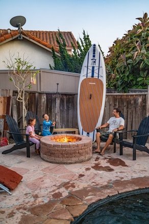 A family scene in a backyard features a man and two children seated around a stone fire pit. The children are sitting on Adirondack chairs, with one holding a roasting stick. Leaned against the wooden fence is a large paddleboard. The setting includes a small tree, a satellite dish on the rooftop of a nearby house, and lush greenery surrounding the area.