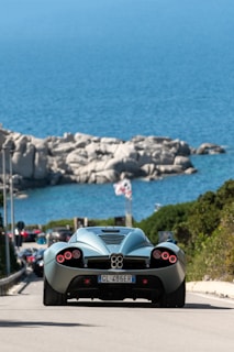 A sleek, futuristic sports car drives along a coastal road, with a clear blue ocean and large rocky formations in the background. The car's distinctive taillights and aerodynamic design dominate the foreground, while the bright sunlight highlights the scene. Several vehicles can be seen behind the car, and a flag is visible near the shoreline.
