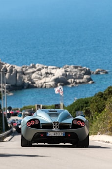 A sleek, futuristic sports car drives along a coastal road, with a clear blue ocean and large rocky formations in the background. The car's distinctive taillights and aerodynamic design dominate the foreground, while the bright sunlight highlights the scene. Several vehicles can be seen behind the car, and a flag is visible near the shoreline.