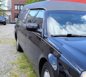 A sleek black hearse parked on a gravel area, with reflections of clouds on its clean, polished surface. The surroundings include some grass patches, a brick building, and another car in the background.