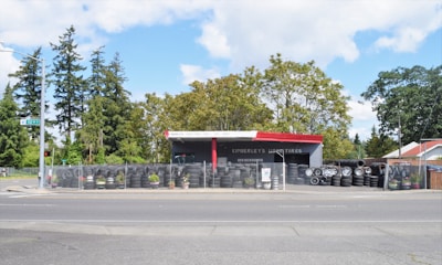 A small tire shop named Kimberley's Used Tires is visible, located by a roadside. The shop has a red and gray exterior with several tires stacked and lined up outside. A chain-link fence surrounds the shop, and there are some potted plants along the fence. The surrounding area includes trees and a clear sky in the background.