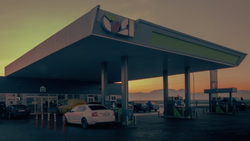A gas station with a canopy and several fueling pumps. Multiple cars are parked near the pumps and possibly waiting for fuel. The scene is set during sunset or sunrise, casting an orange hue across the landscape. The structure is modern, with columns supporting the roof, and there are signs indicating different services. A spacious forecourt with a building in the background possibly houses a convenience store.