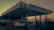 A gas station with a canopy and several fueling pumps. Multiple cars are parked near the pumps and possibly waiting for fuel. The scene is set during sunset or sunrise, casting an orange hue across the landscape. The structure is modern, with columns supporting the roof, and there are signs indicating different services. A spacious forecourt with a building in the background possibly houses a convenience store.