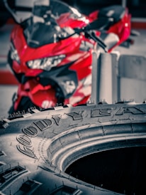 A close-up view of a Goodyear tire with fine details of the tread and brand name visible. Behind the tire, a red motorcycle is out of focus, adding a vibrant pop of color to the scene.
