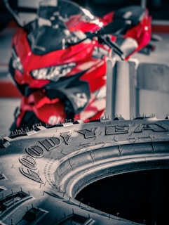 A close-up view of a Goodyear tire with fine details of the tread and brand name visible. Behind the tire, a red motorcycle is out of focus, adding a vibrant pop of color to the scene.