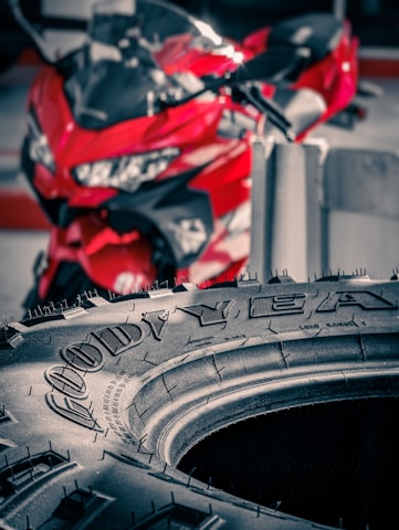 A close-up view of a Goodyear tire with fine details of the tread and brand name visible. Behind the tire, a red motorcycle is out of focus, adding a vibrant pop of color to the scene.