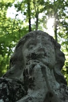 A weathered stone statue of a figure with hands clasped in prayer stands against a backdrop of lush green trees. Sunlight filters through the canopy, creating a starburst effect above the statue's head, adding a divine aura to the scene.