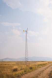 A tall wind turbine stands in an open field with grasslands stretching out towards distant hills. The sky is partly cloudy with light blue hues, adding to the tranquil setting. A dirt path runs parallel to the scene, indicating a rural location.