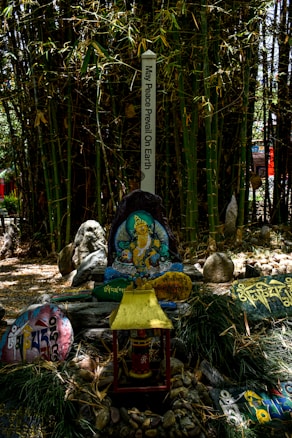 A spiritual garden setting featuring a peace pole with the inscription 'May Peace Prevail On Earth'. Surrounding the pole are rocks and slabs with colorful inscriptions and illustrations, including a central depiction of a deity or spiritual figure. The background is dense with bamboo plants, creating a serene and lush environment.