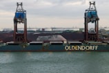A large cargo ship named Oldendorff is docked at an industrial port. Two towering cranes stand above the ship, indicating loading or unloading activities. In the background, there are large piles of materials, possibly coal or gravel, with industrial buildings and storage tanks visible under an overcast sky.