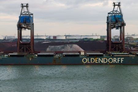A large cargo ship named Oldendorff is docked at an industrial port. Two towering cranes stand above the ship, indicating loading or unloading activities. In the background, there are large piles of materials, possibly coal or gravel, with industrial buildings and storage tanks visible under an overcast sky.