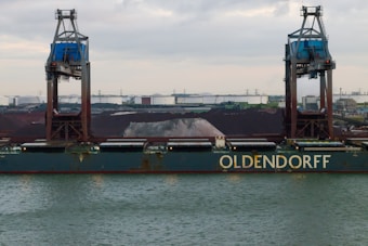 A large cargo ship named Oldendorff is docked at an industrial port. Two towering cranes stand above the ship, indicating loading or unloading activities. In the background, there are large piles of materials, possibly coal or gravel, with industrial buildings and storage tanks visible under an overcast sky.