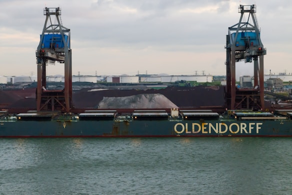 A large cargo ship named Oldendorff is docked at an industrial port. Two towering cranes stand above the ship, indicating loading or unloading activities. In the background, there are large piles of materials, possibly coal or gravel, with industrial buildings and storage tanks visible under an overcast sky.
