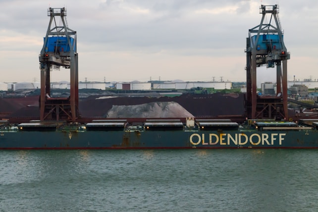 A large cargo ship named Oldendorff is docked at an industrial port. Two towering cranes stand above the ship, indicating loading or unloading activities. In the background, there are large piles of materials, possibly coal or gravel, with industrial buildings and storage tanks visible under an overcast sky.
