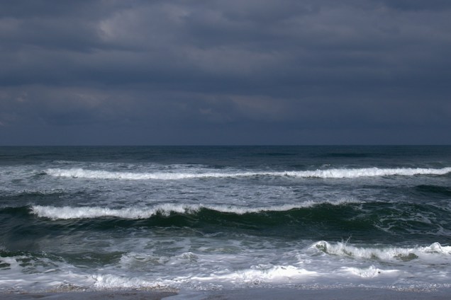 Dark, cloud-filled sky over a vast, turbulent ocean with waves crashing towards the shore.