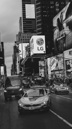 A bustling urban scene with various cars, including a sleek BMW and a Mercedes van, driving through a busy city street lined with towering buildings displaying illuminated advertisements. Crowds of people populate the sidewalks, contributing to the dynamic atmosphere of the city.