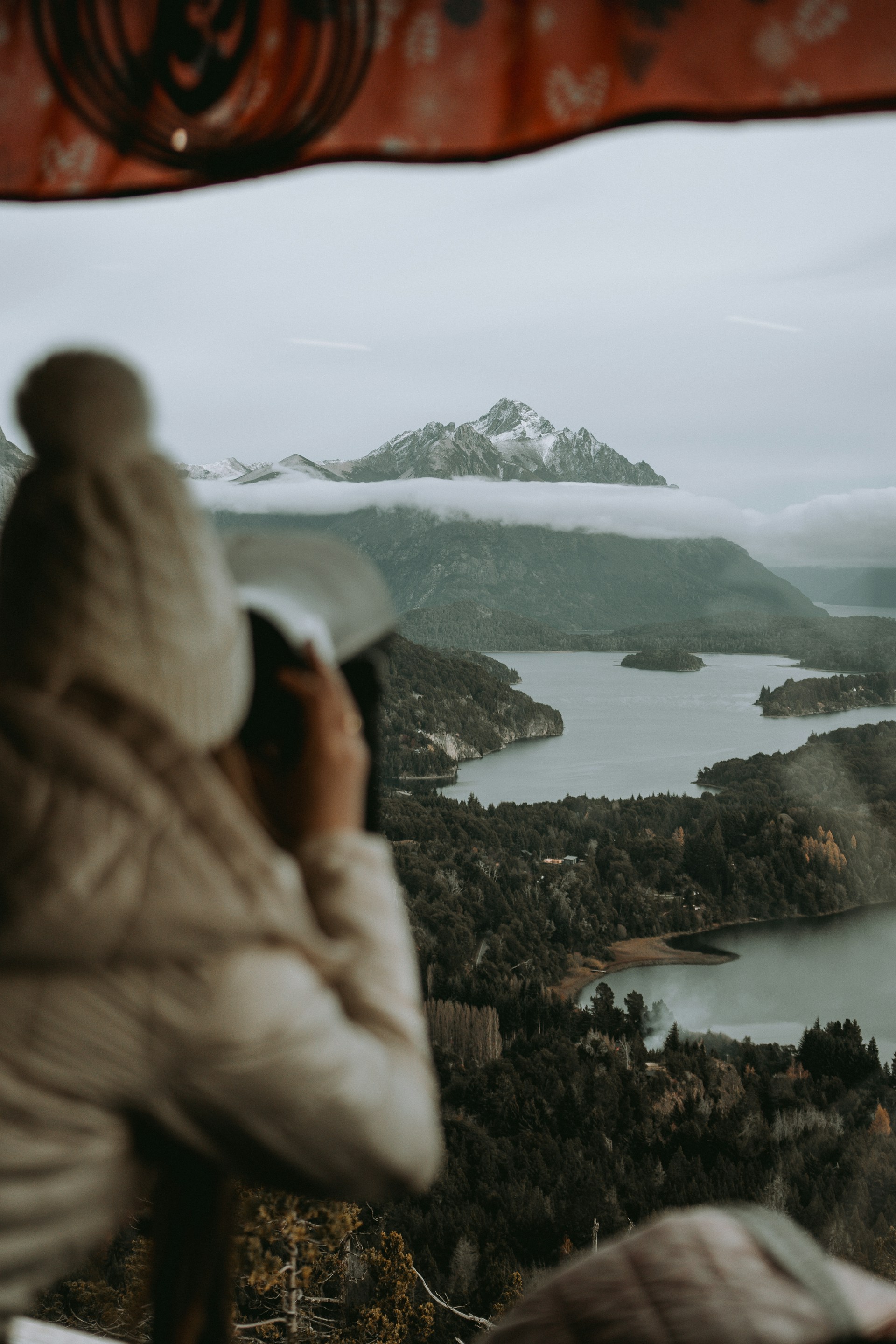 A person wearing a knit hat and jacket observes and photographs a scenic landscape of a tranquil lake and forested hills, with mountains shrouded in misty clouds in the background. A patterned fabric or structure frames the top of the image.