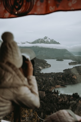 A person wearing a knit hat and jacket observes and photographs a scenic landscape of a tranquil lake and forested hills, with mountains shrouded in misty clouds in the background. A patterned fabric or structure frames the top of the image.