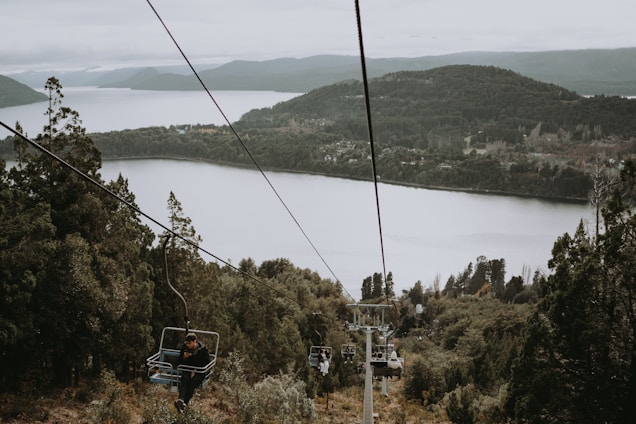 A serene landscape featuring a large lake surrounded by forested hills. Several cable cars are visible, with people seated as they ascend or descend amidst the trees. The scenery conveys a sense of tranquility and connection with nature.