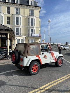 A white Jeep with red accents and a Jurassic Park logo is parked on a city street. The vehicle has distinctive red rims and a brown soft top. It is near a large multi-story yellow building with many windows, under a partly cloudy blue sky. Several street lamps and a motorcycle are visible in the background, along with a few pedestrians and cars.
