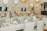 A beautifully arranged table setting in an elegant venue, featuring white tablecloths, beige napkins, and floral centerpieces in sleek vases. The background includes white balloons and soft lighting, creating a sophisticated ambiance.