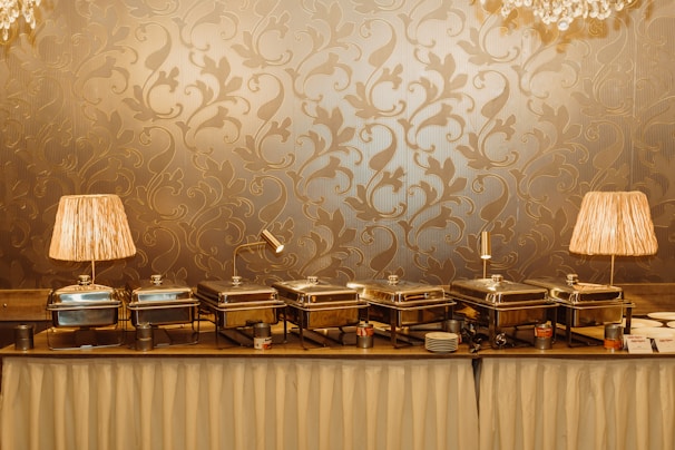 A buffet setup with several silver chafing dishes neatly arranged on a table covered with a white cloth. Two decorative lamps with pleated beige shades illuminate the scene from either end. The background features an ornate, gold-patterned wallpaper, adding a touch of elegance.