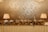 A buffet setup with several silver chafing dishes neatly arranged on a table covered with a white cloth. Two decorative lamps with pleated beige shades illuminate the scene from either end. The background features an ornate, gold-patterned wallpaper, adding a touch of elegance.