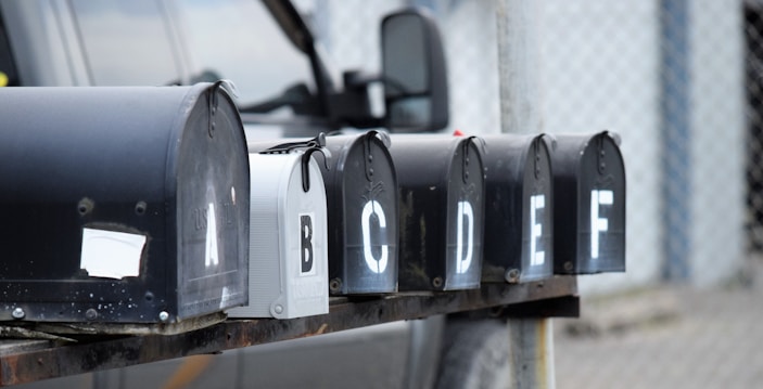 A row of six black mailboxes is mounted on a metal beam. Each mailbox is labeled with a white letter from A to F. The background includes part of a vehicle and a chain-link fence.