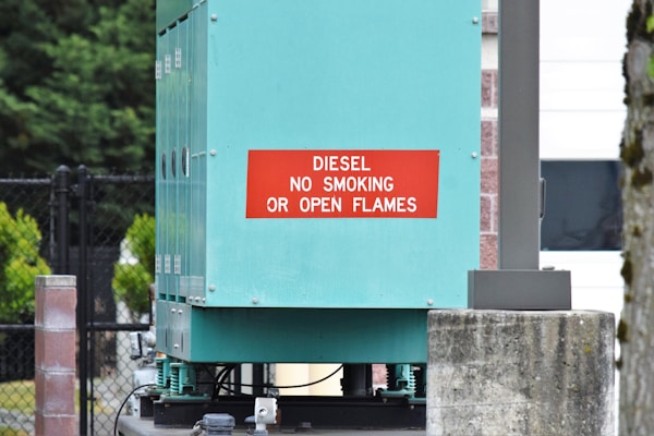 The image displays an industrial setup with a teal-colored equipment box featuring a red sign that reads 'Diesel No Smoking or Open Flames'. Surrounding the box is a black chain-link fence, brick pillars, and some greenery is visible in the background.