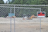 A chain-link fence encloses an area with construction vehicles in the background. A prominent 'No Trespassing' sign is attached to the fence. Dense trees form a lush green backdrop.