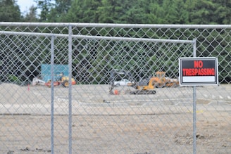 A chain-link fence encloses an area with construction vehicles in the background. A prominent 'No Trespassing' sign is attached to the fence. Dense trees form a lush green backdrop.
