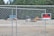 A chain-link fence encloses an area with construction vehicles in the background. A prominent 'No Trespassing' sign is attached to the fence. Dense trees form a lush green backdrop.