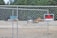 A chain-link fence encloses an area with construction vehicles in the background. A prominent 'No Trespassing' sign is attached to the fence. Dense trees form a lush green backdrop.