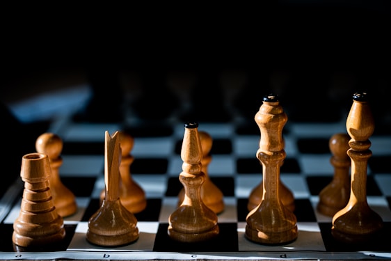 A close-up view of wooden chess pieces neatly arranged on a chessboard. The pieces include a bishop, knight, rook, and queen. The lighting creates a dramatic effect with shadows and highlights on the pieces, emphasizing their polished wooden texture.