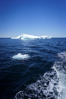 A vast ocean stretches toward a distant iceberg under a clear blue sky. The iceberg is large and shines with white and light blue hues against the deep blue of the water. In the foreground, a smaller piece of ice floats, and the water is slightly frothy, suggesting movement.