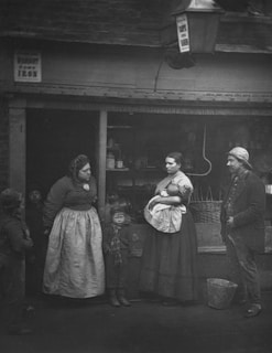 A group of individuals, including two women, a man, and two children, stand in front of a rustic building. One woman cradles a baby, while another stands with hands on hips. The attire and setting suggest a historical period, possibly late 19th to early 20th century. The expressions are serious and the environment seems modest.