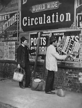 Two men are seen putting up posters on a wall covered with various advertisements. One man is pasting a poster while another stands with a bucket and holds some tools. Both are dressed in old-fashioned work attire, including hats. The wall is heavily covered in advertisements for a variety of products and services.