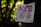 A sign with red text indicating 'POSTED PRIVATE PROPERTY' is attached to a tree in a wooded area. The text further states that hunting, fishing, trapping, or trespassing for any purpose is strictly forbidden and violators will be prosecuted. The sign shows some weathering and signs of dirt.