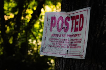 A sign with red text indicating 'POSTED PRIVATE PROPERTY' is attached to a tree in a wooded area. The text further states that hunting, fishing, trapping, or trespassing for any purpose is strictly forbidden and violators will be prosecuted. The sign shows some weathering and signs of dirt.