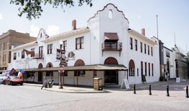 A large white historic building with Spanish-style architecture on a cobblestone street. The facade features wooden awnings, balconies, and a sign reading 'Fincher's Western Frontier Western Store.' Flags, including a Texas flag, are displayed at one end of the building. A parked motorcycle and car can be seen in front, while a 'No Entry' sign is positioned nearby. The scene is calm with few pedestrians.