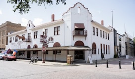 A large white historic building with Spanish-style architecture on a cobblestone street. The facade features wooden awnings, balconies, and a sign reading 'Fincher's Western Frontier Western Store.' Flags, including a Texas flag, are displayed at one end of the building. A parked motorcycle and car can be seen in front, while a 'No Entry' sign is positioned nearby. The scene is calm with few pedestrians.