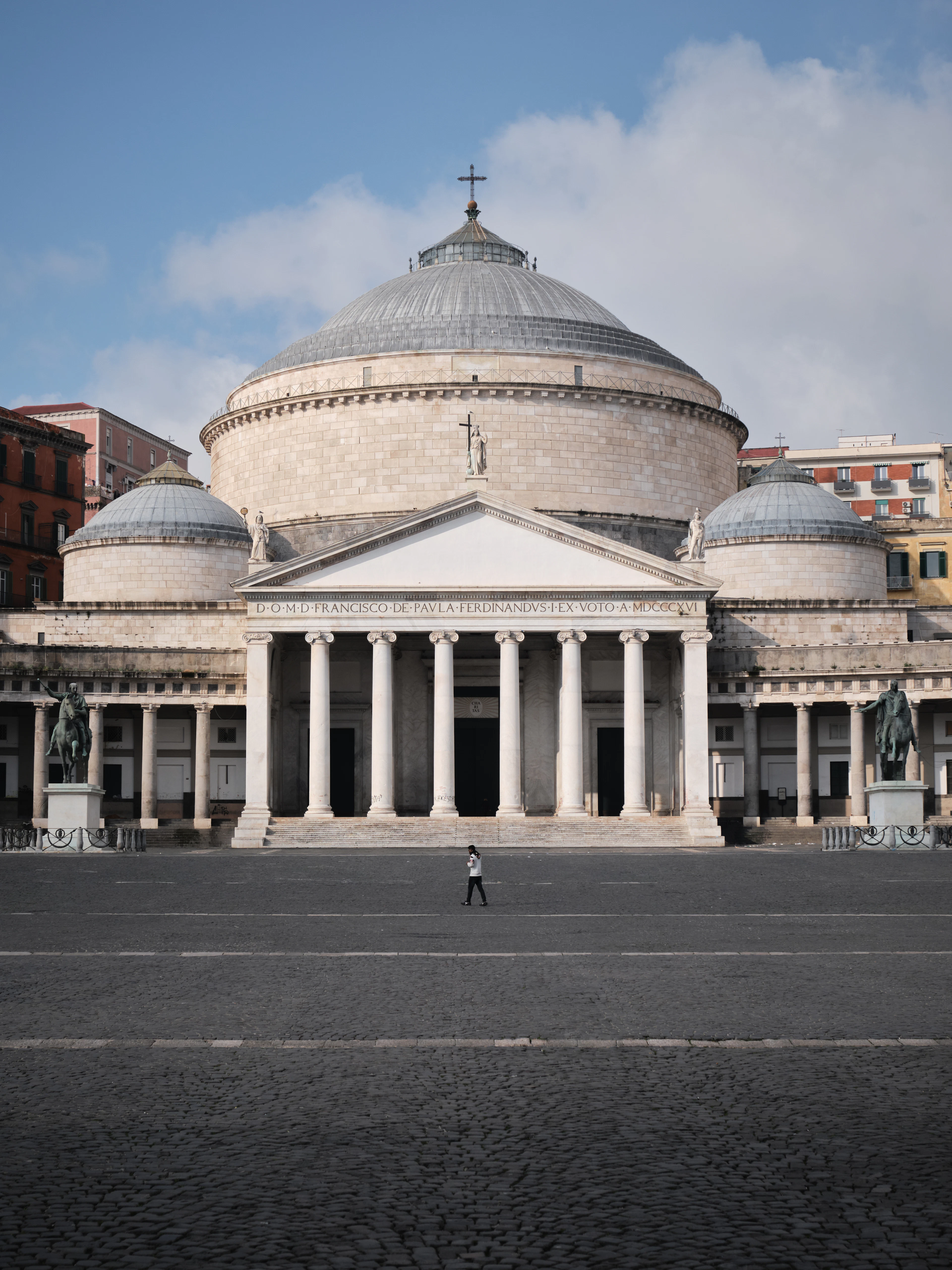 The domed neoclassical church of San Francesco di Paola behind its colonnaded portico and equestrian statues, seen across the empty grey cobbles of Piazza del Plebiscito with one figure crossing.