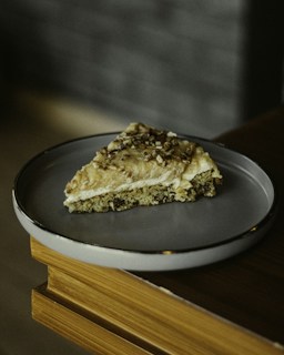 A piece of cake with a nutty topping rests on a simple, round plate. The cake has a light brown color and appears to be moist, with visible nuts or seeds embedded on the surface. It is placed on a wooden surface with a muted backdrop, suggesting a cozy indoor setting.