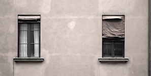 Two windows are set in a plain, gray concrete wall. The window on the left has partially open blinds with visible white curtains, while the window on the right is covered with a wrinkled, closed blind. The facade appears slightly weathered, creating a minimalistic and somewhat industrial look.