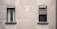 Two windows are set in a plain, gray concrete wall. The window on the left has partially open blinds with visible white curtains, while the window on the right is covered with a wrinkled, closed blind. The facade appears slightly weathered, creating a minimalistic and somewhat industrial look.